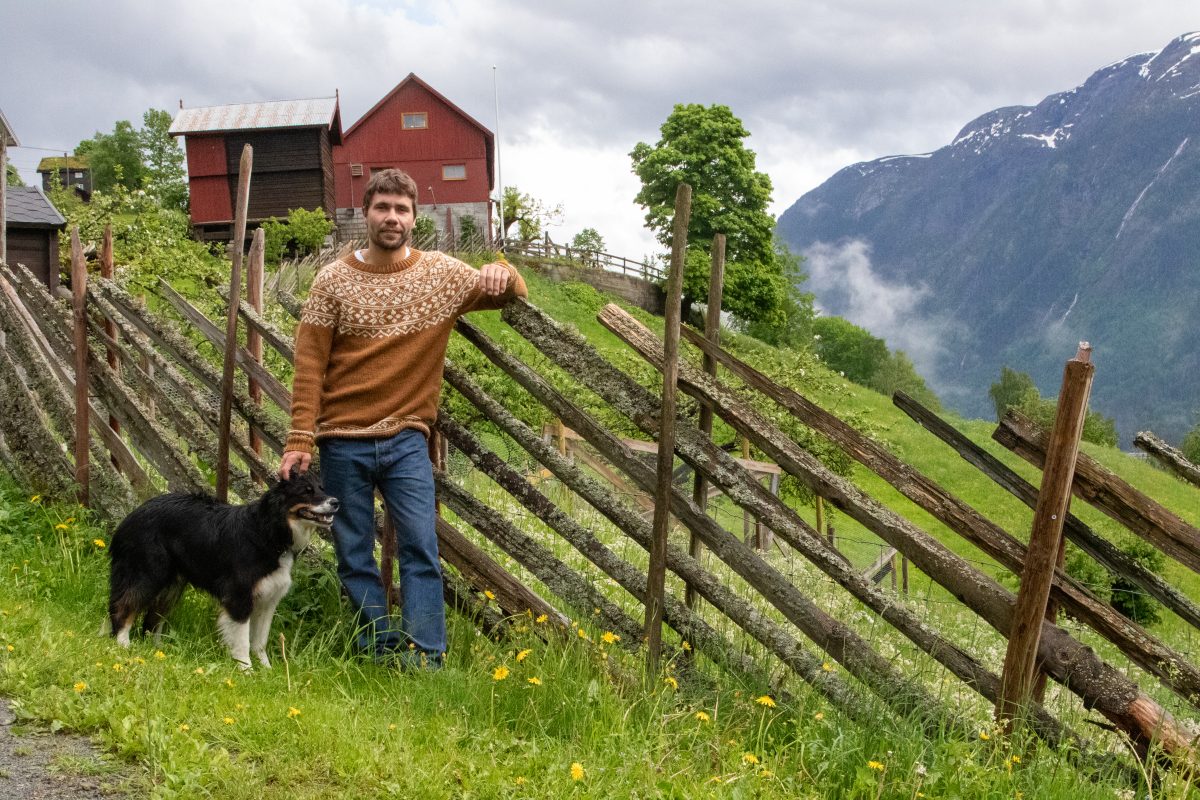 Gunleik Mæland fra Rørtveit Gard står ved et skigardsgjerde sammen med hunden sin, med gårdstun og fjellandskap i bakgrunnen.