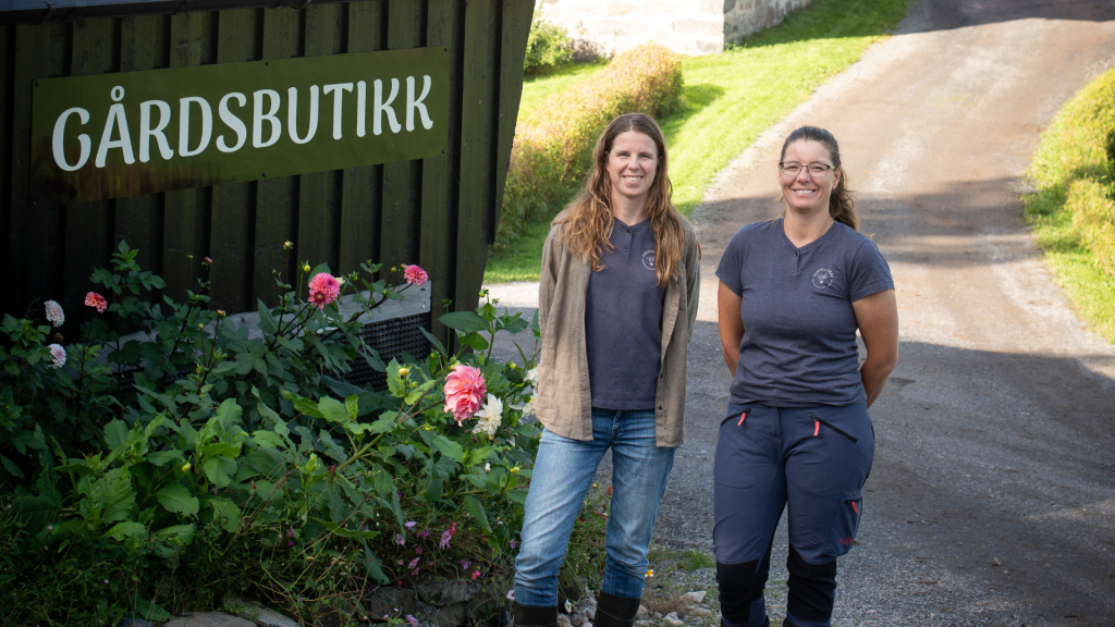 To personer står utenfor en norsk gårdsbutikk med skiltet «Gårdsbutikk», omgitt av blomster, gårdstun og landlige omgivelser.