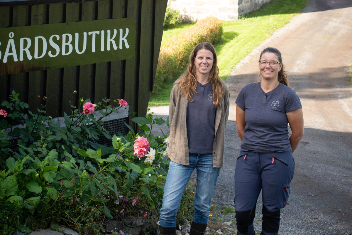 To personer står utenfor en norsk gårdsbutikk med skiltet «Gårdsbutikk», omgitt av blomster, gårdstun og landlige omgivelser.