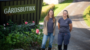 To personer står utenfor en norsk gårdsbutikk med skiltet «Gårdsbutikk», omgitt av blomster, gårdstun og landlige omgivelser.