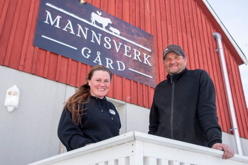 Driverne ved Mannsverk gård står foran den røde gårdsbygningen med skiltet «Mannsverk gård».